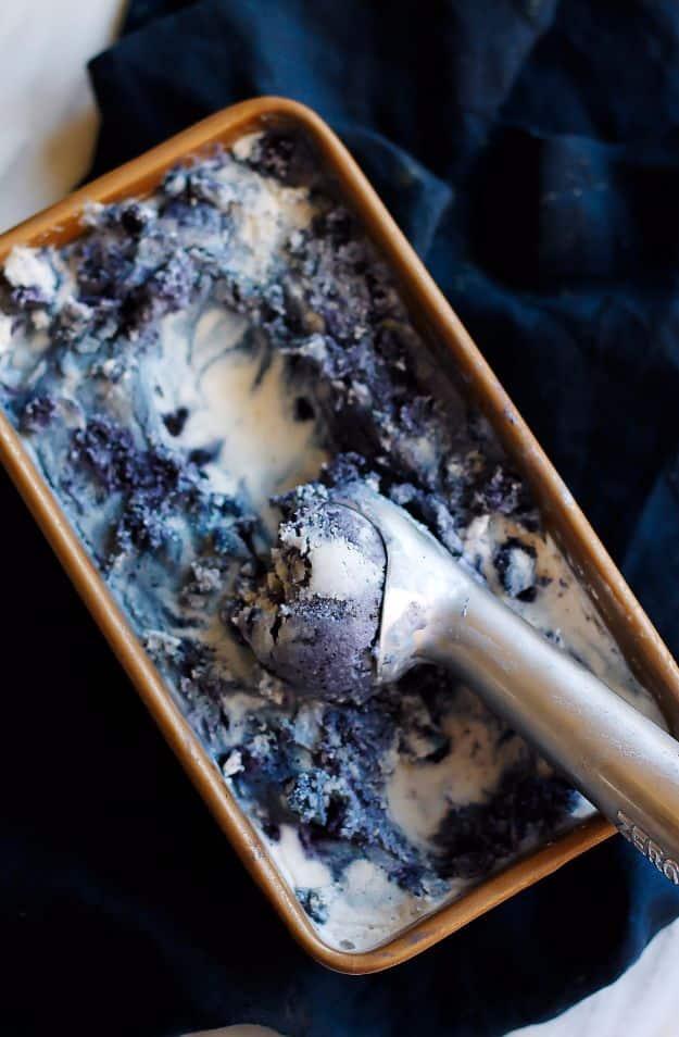 Loaf tin of coconut ice cream ribboned with dark blueberries, a metal scoop resting in it on navy cloth