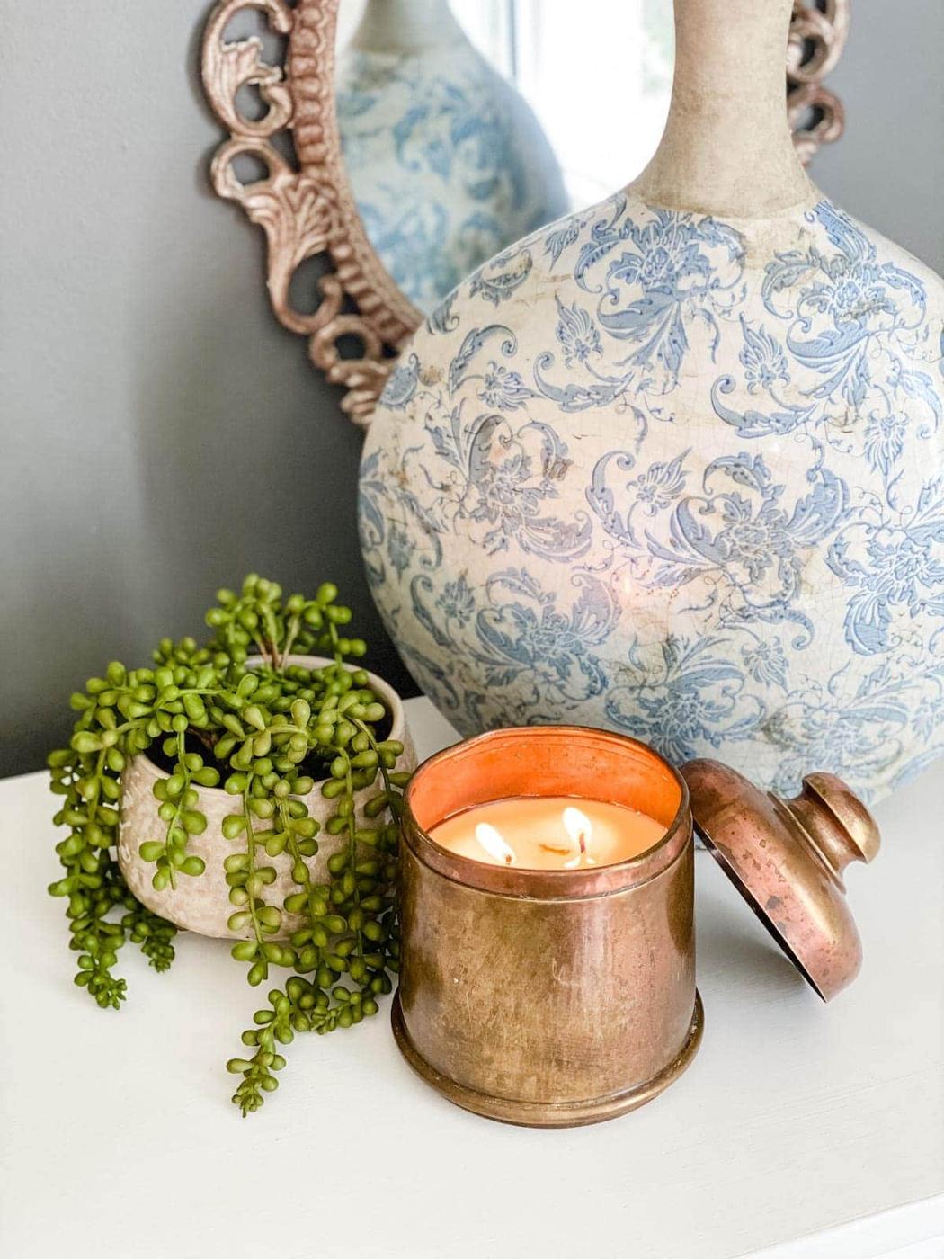 A lit two wick candle in a thrifted brass canister with its lid beside it, next to a small potted string of pearls plant