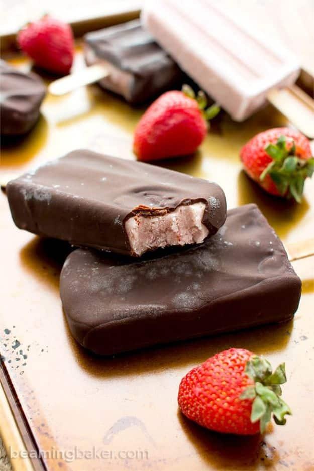 Chocolate covered strawberry ice cream bars on a wooden board, one bitten to show the pink strawberry center