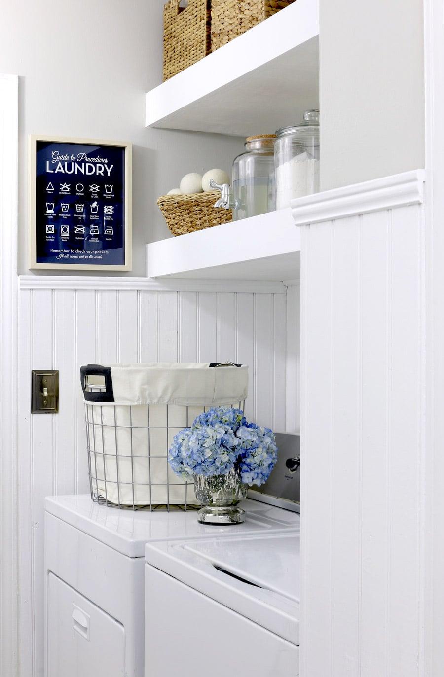 White floating shelves, two thick white shelves above a washer and dryer holding baskets and glass storage jars