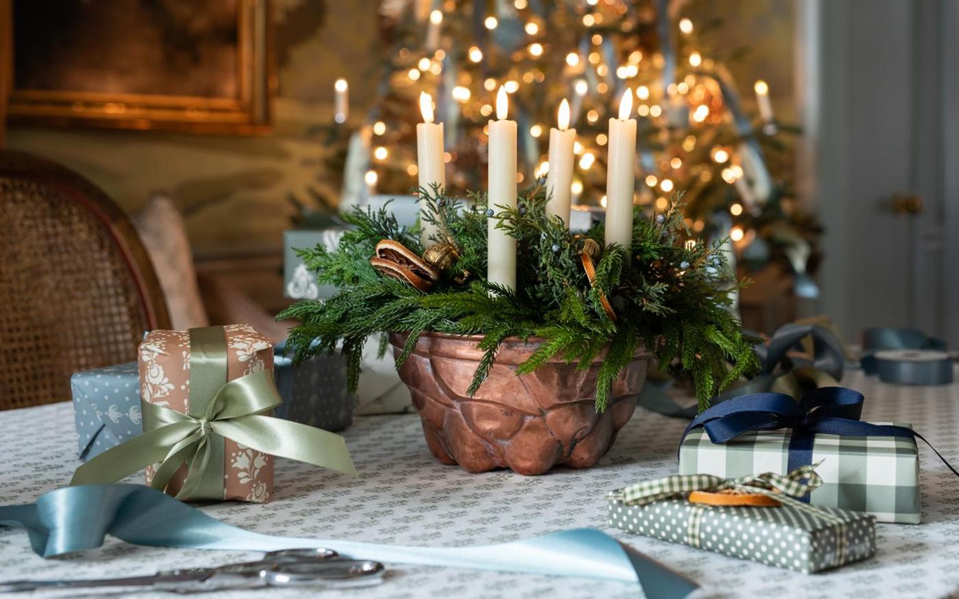 An antique copper mold holding four cream taper candles among faux juniper sprigs and brass sleigh bells on a wood table