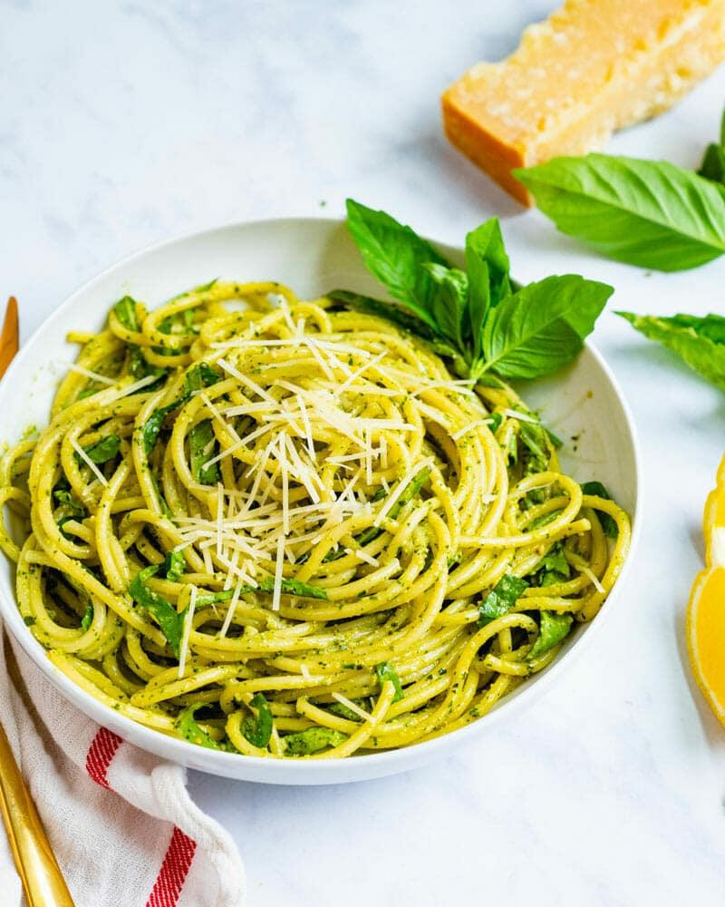 Basil pesto pasta, bright green spaghetti in a white bowl beside fresh basil leaves and a wedge of parmesan