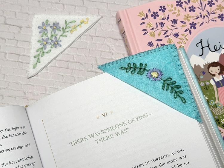 Two triangle felt corner bookmarks embroidered with small flowers resting on an open book and a stack of books