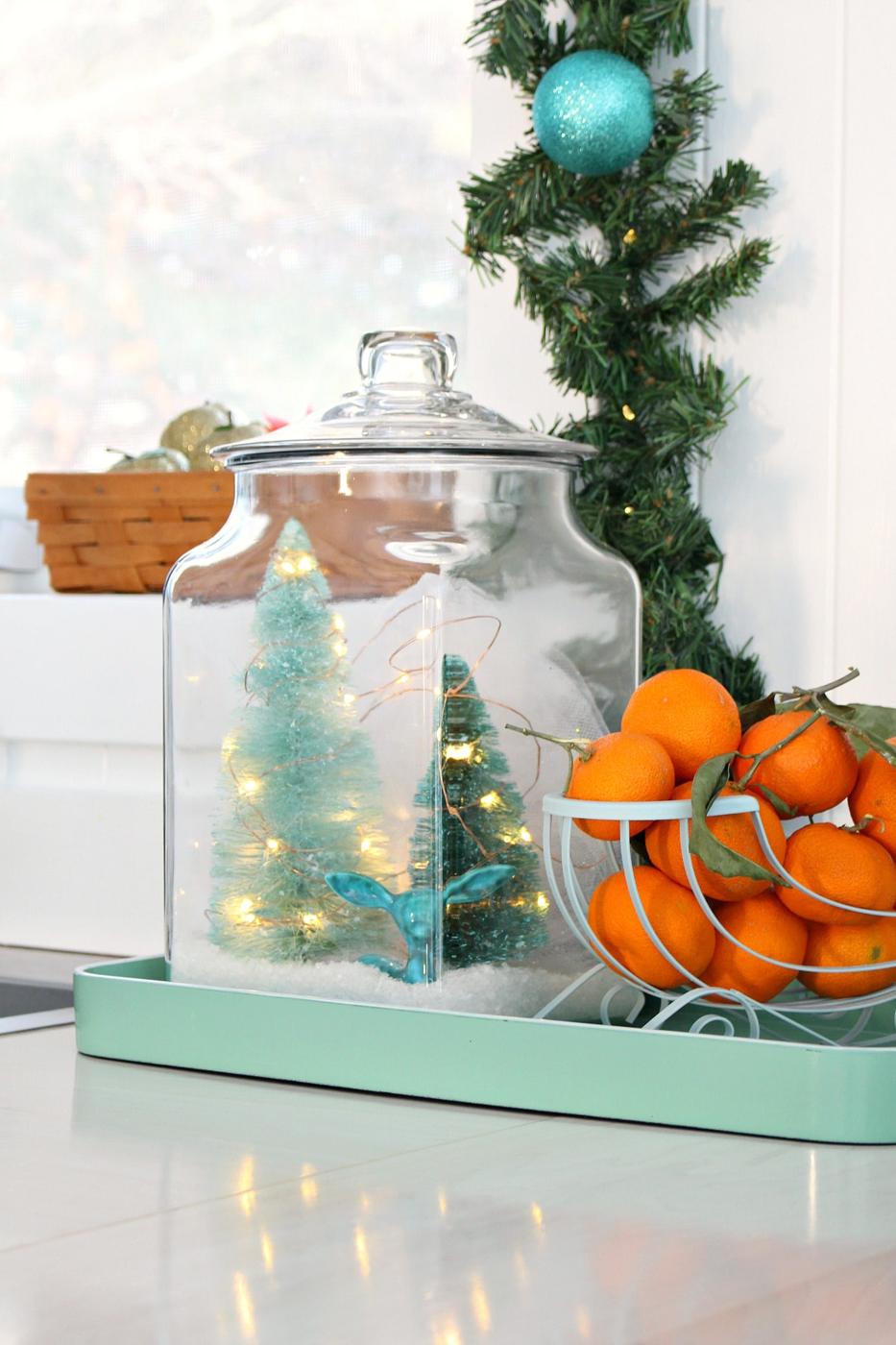 Large glass storage jar holding two aqua bottle brush trees, a teal ceramic deer, faux snow and wire lights on a mint tray