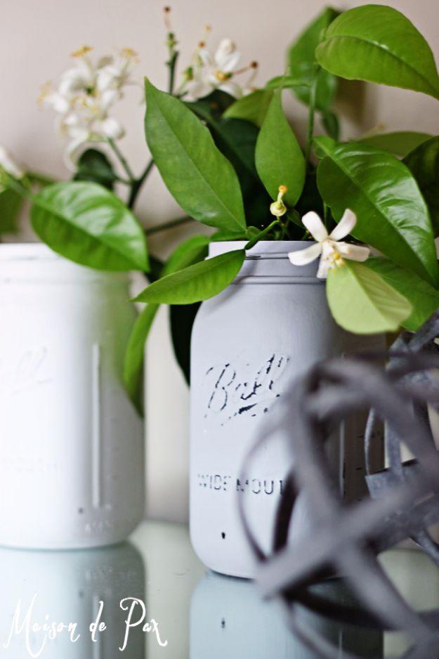 Two chalk painted mason jars in white and soft gray holding white blossoms and green leaves on a counter