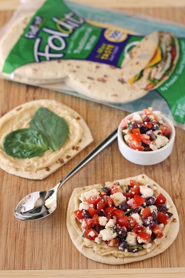 Greek snack flats with hummus, spinach, tomato, olives and feta on flatbread rounds beside the package