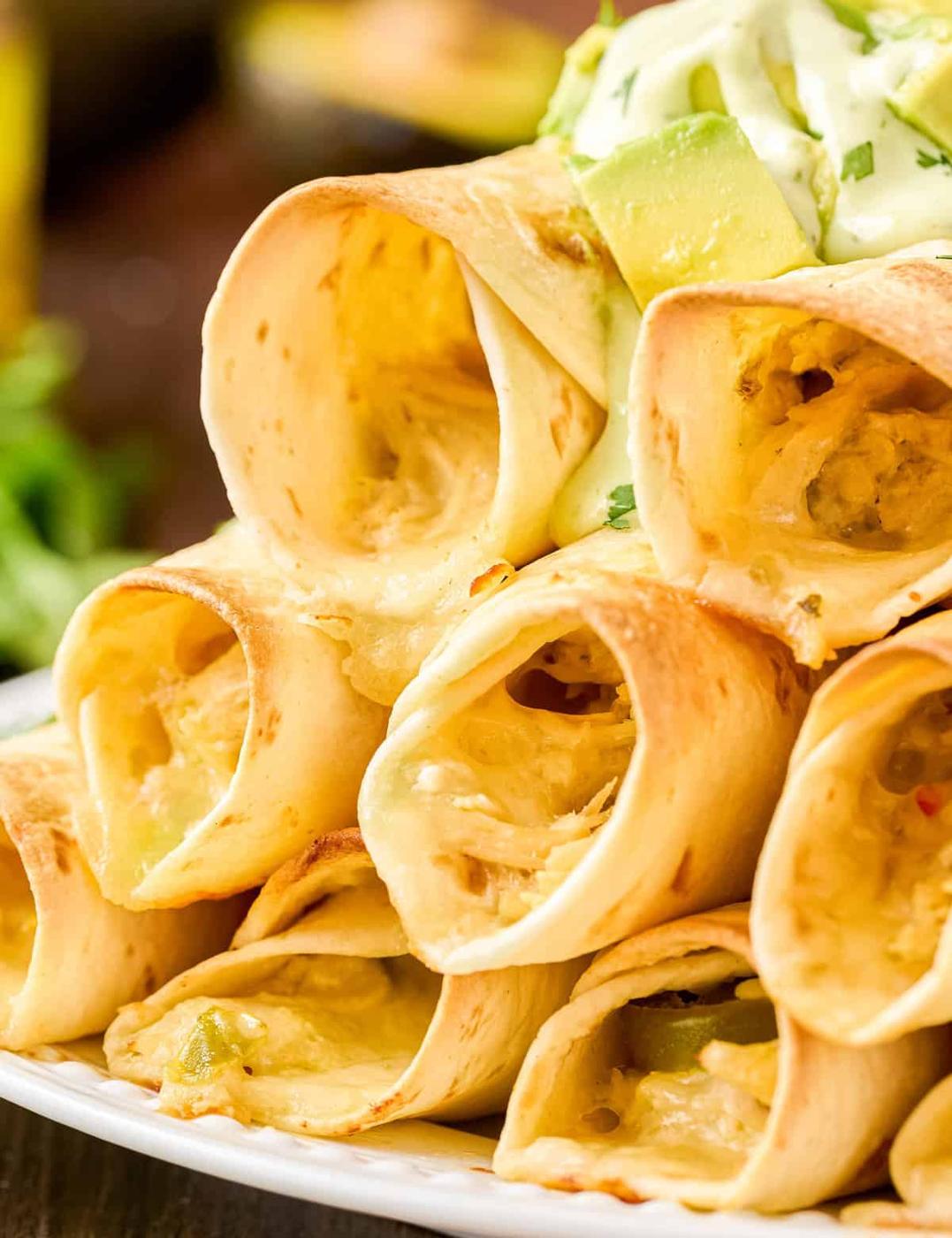 Baked chicken taquitos stacked on a plate with melted cheese inside and chopped avocado over the top
