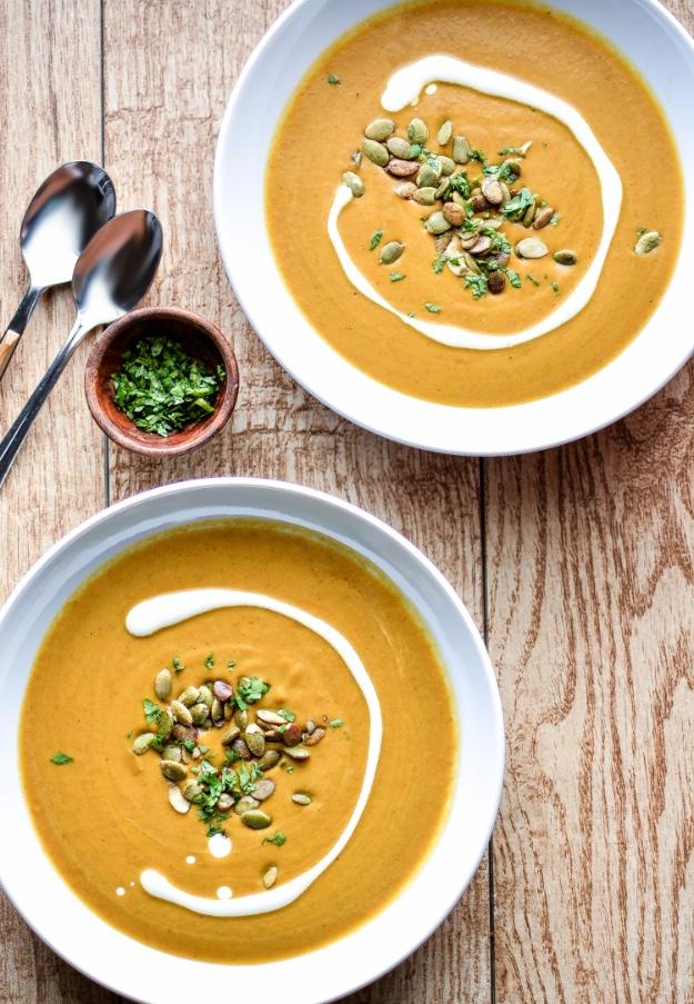 Creamy orange pumpkin soup with cream swirls, seeds, herbs, spoons and a rustic wood table in an overhead view