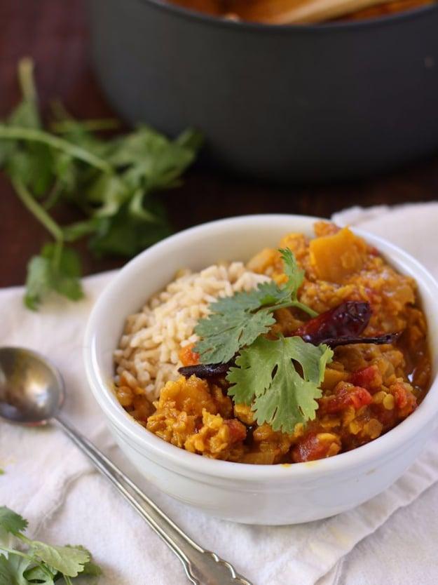 Red lentil pumpkin dal in a white bowl with brown rice beside it, cilantro and two dried red chilies over the top