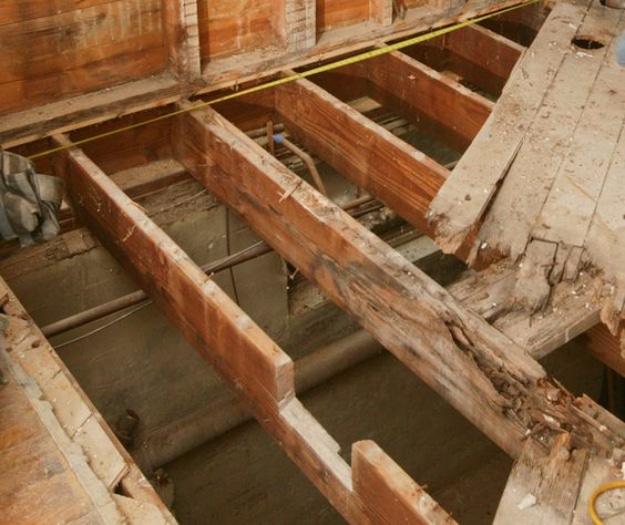 DIY home repair: cut and notched floor joists seen from above with the subfloor lifted, pipes running beneath