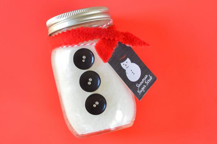 A pint jar of white sugar scrub dressed as a snowman with three black buttons, a red felt scarf and a printed snowman tag