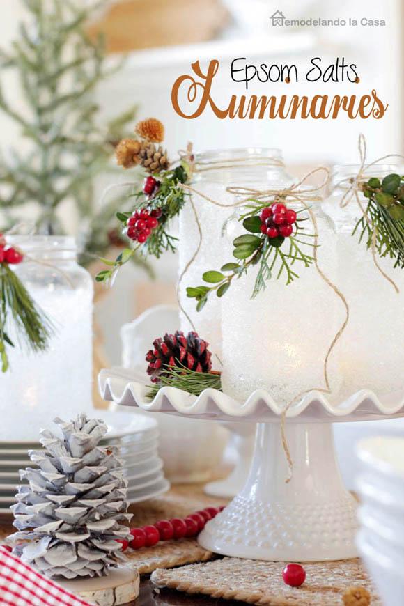 Epsom salt frosted jar luminaries with twine, greenery and red berries glowing on a white cake stand beside a white pine cone