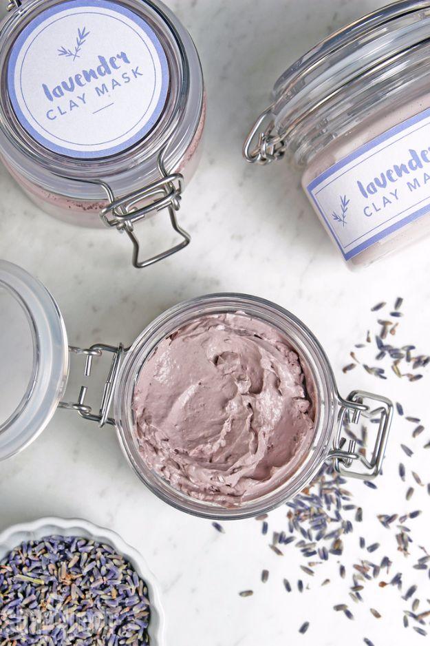 Open glass jar of pale purple lavender clay face mask with labeled jars and dried buds on white marble