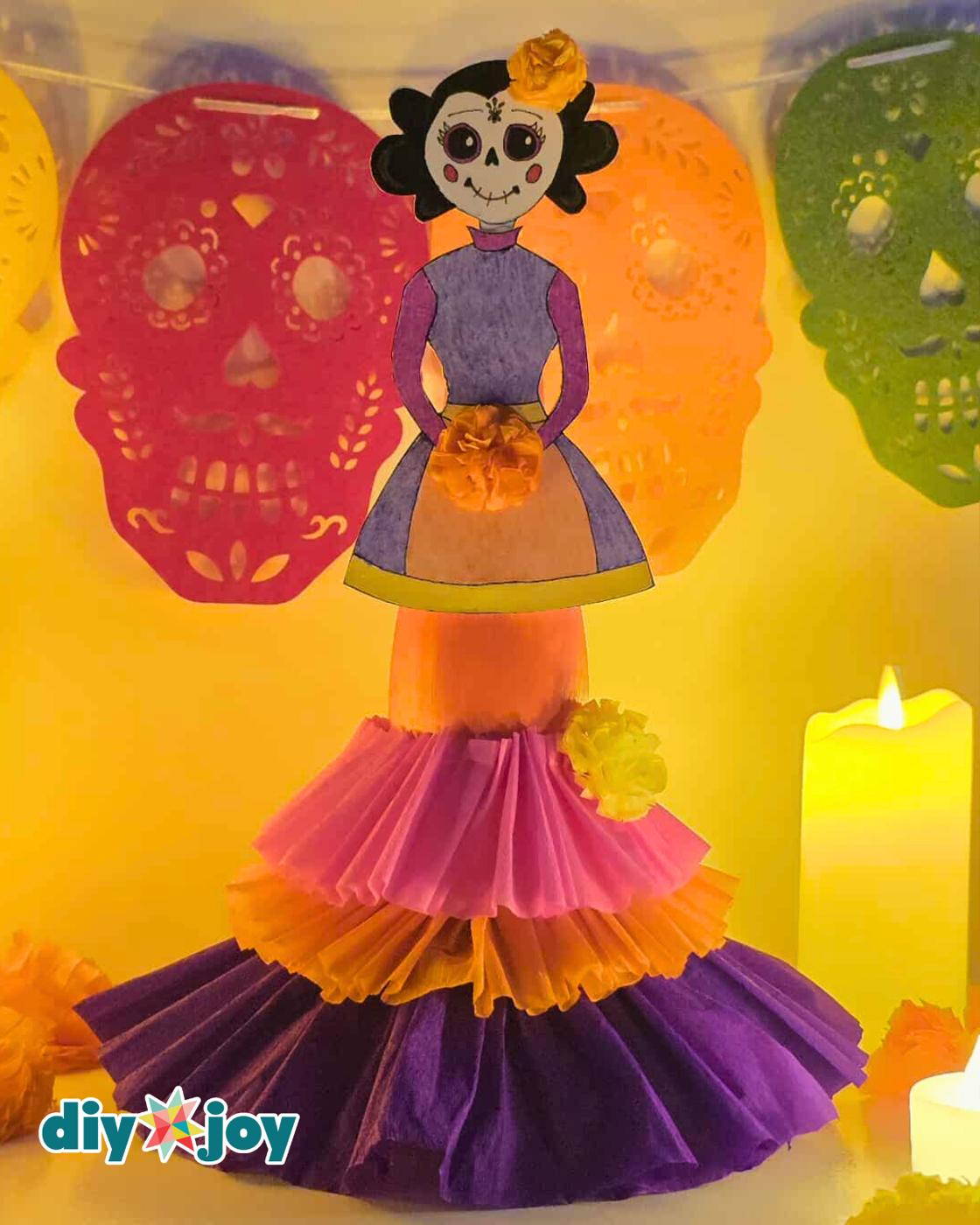 DIY Ofrenda - the full-length finished La Catrina in amber lighting, standing in front of a yellow, red, orange and green papel picado banner with a lit candle at lower right