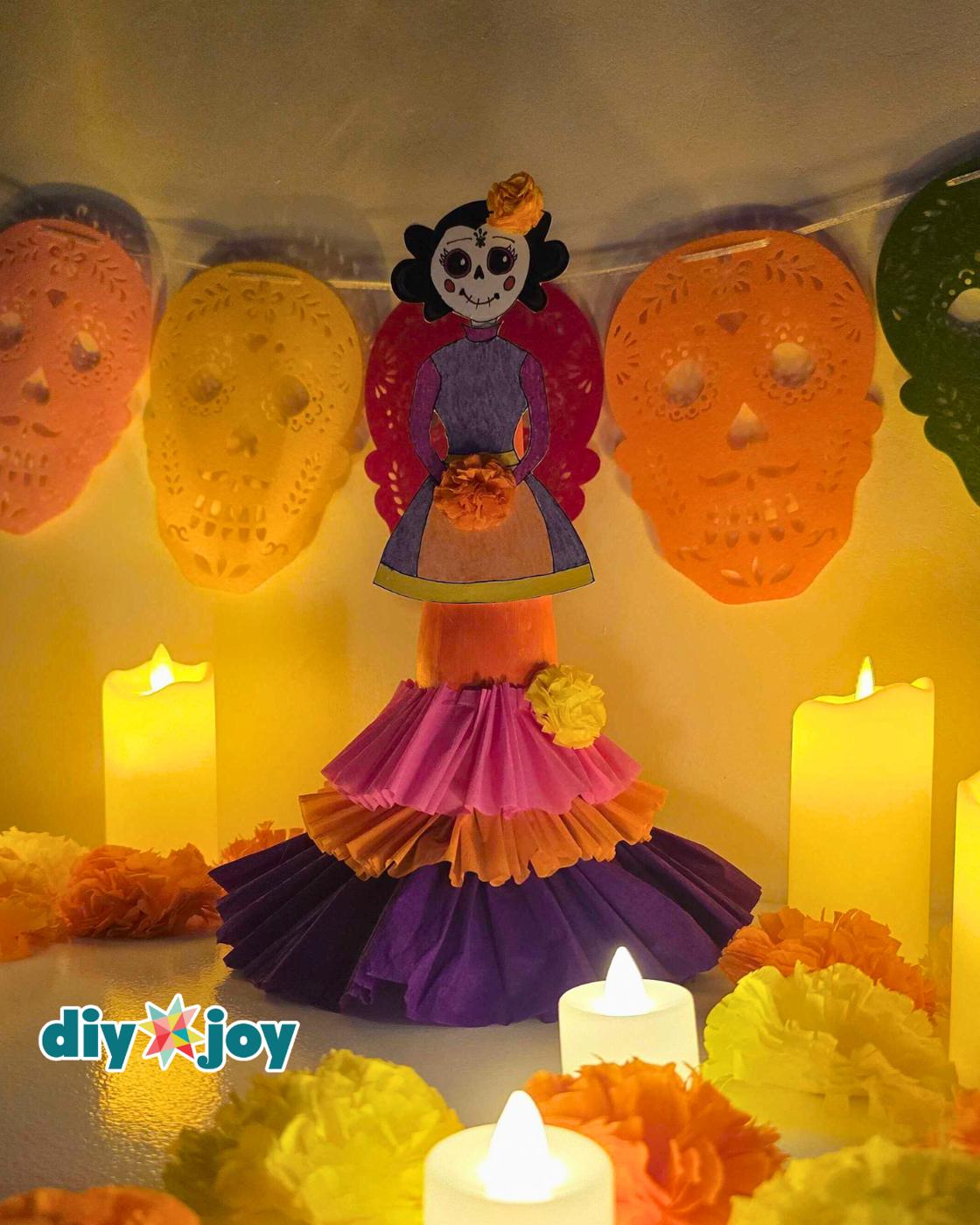 La Catrina - the finished bottle figure photographed indoors under warm light, one lit pillar candle to her left and marigold tissue flowers at her base