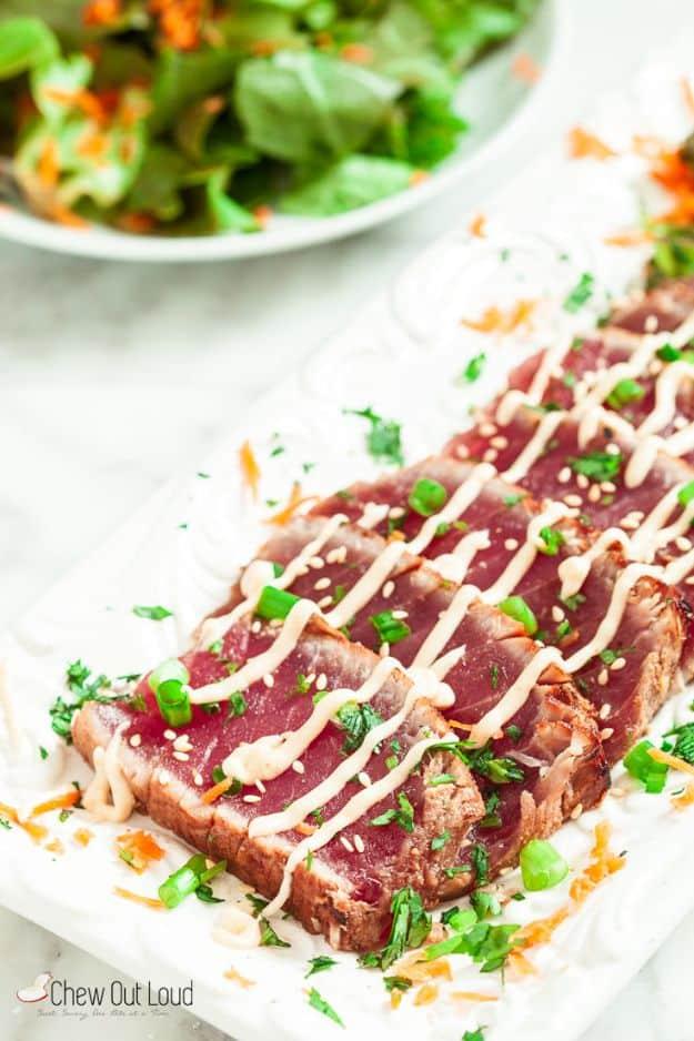 Sliced seared ahi tuna fanned on a white platter with sriracha mayo drizzle and a green salad behind