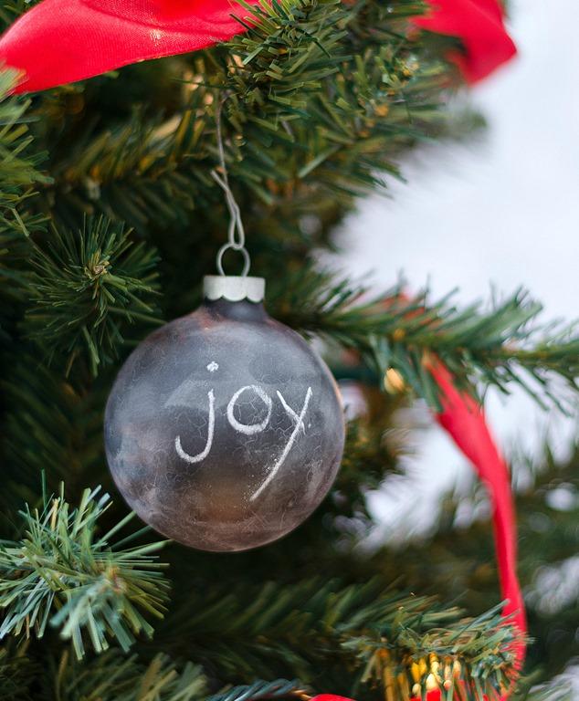A black chalkboard ball ornament with the word joy in chalk hanging on a wire hook in an evergreen tree with red ribbon
