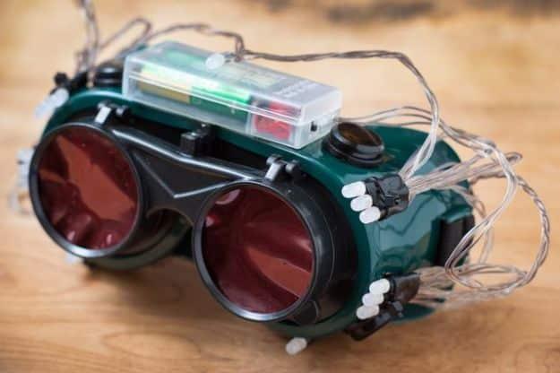 Green goggles with deep red lenses, a clear battery box taped on top and white LEDs wired around the frame