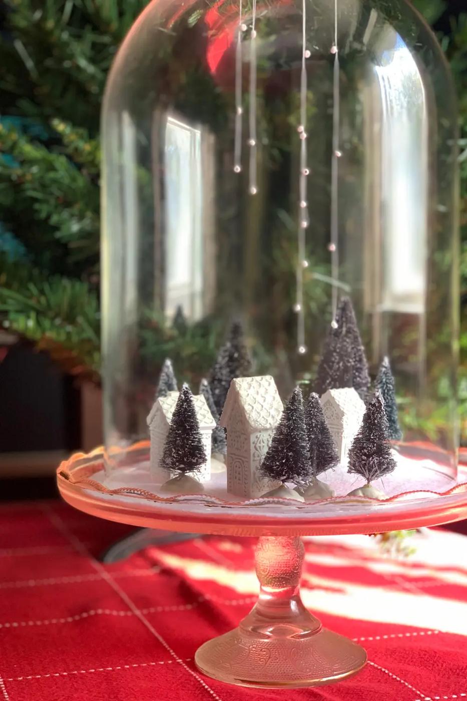 Glass cloche on a cake plate covering a tiny Christmas village of white glittered houses and green sisal trees on felt snow