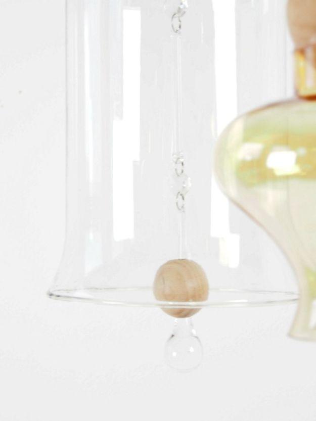 Clear glass bell shapes and a pale wooden bead hanging on a fine chain against a white wall, with an amber glass shape behind