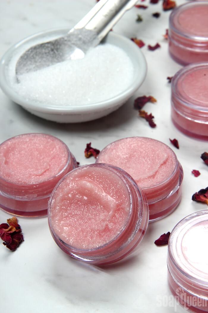 Four small clear pots of pale pink sugar lip scrub beside a bowl of white sugar and scattered dried rose petals