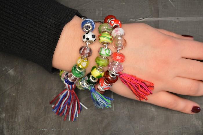 A stack of colorful beaded bangle bracelets with a multicolored floss tassel worn on a wrist against a gray wall