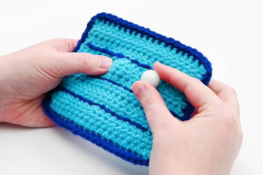 A crocheted blue fidget toy pouch with dark blue maze channels and a white marble inside, held in two hands on white