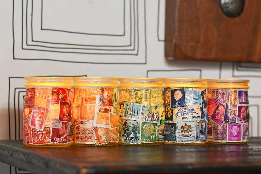 Five small glass jars decoupaged with vintage postage stamps in red, orange, yellow, green and purple glowing on a shelf