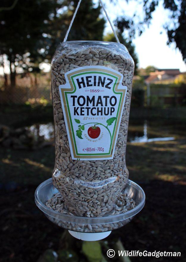 Upside down Heinz tomato ketchup bottle bird feeder filled with sunflower hearts above a clear round catch tray