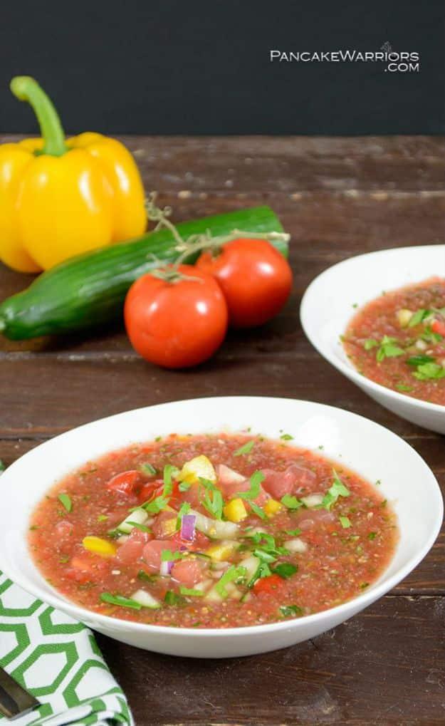 Easy watermelon gazpacho in two white bowls topped with chopped vegetables beside a yellow pepper and tomatoes