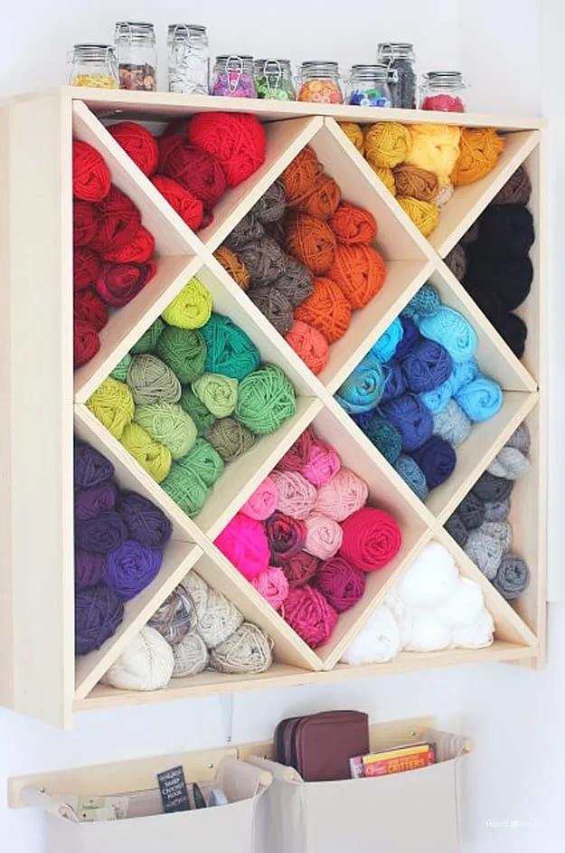 Wall mounted wine rack with diamond cubbies filled with yarn skeins sorted by color, canvas bins hanging below