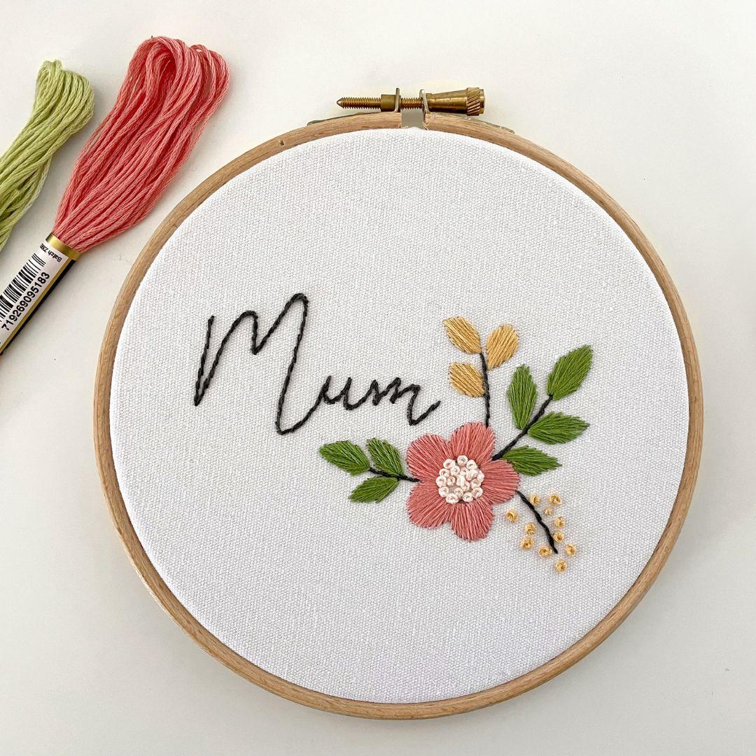 Wooden embroidery hoop with the word Mum stitched in gray beside a coral flower and green leaves, with floss skeins nearby