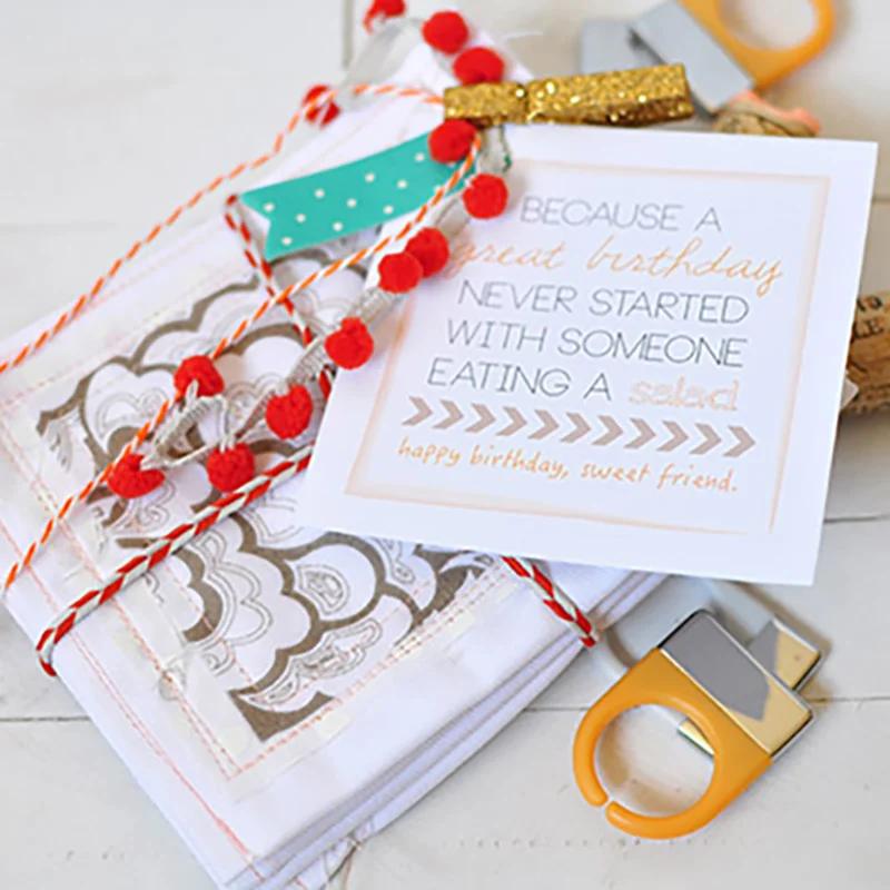 White cloth napkins stamped with a gray scalloped pattern, tied with red pom pom trim and twine beside a birthday card