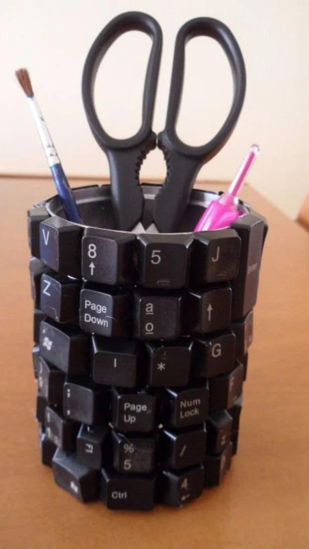 Pencil cup covered in black keyboard keys holding scissors, a paintbrush and colored pens on a white surface