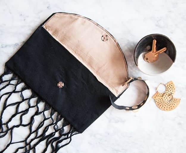 A suede fringed clutch in tan, fringe hanging loose, photographed on a dark table beside a gold bracelet.