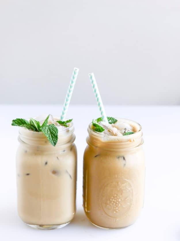 Two mason jars of creamy iced coffee with striped paper straws and fresh mint sprigs on a white counter