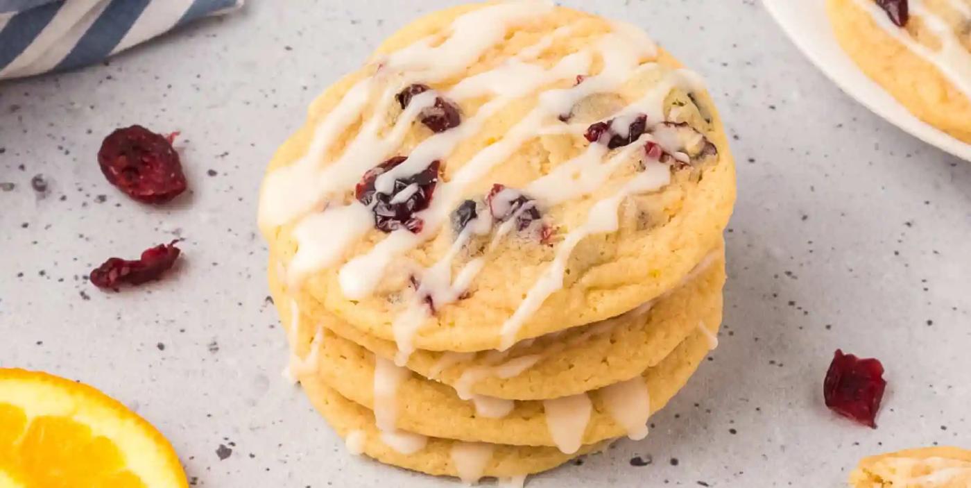A stack of soft orange cranberry cookies with dried cranberries and a white orange glaze drizzle on a gray surface