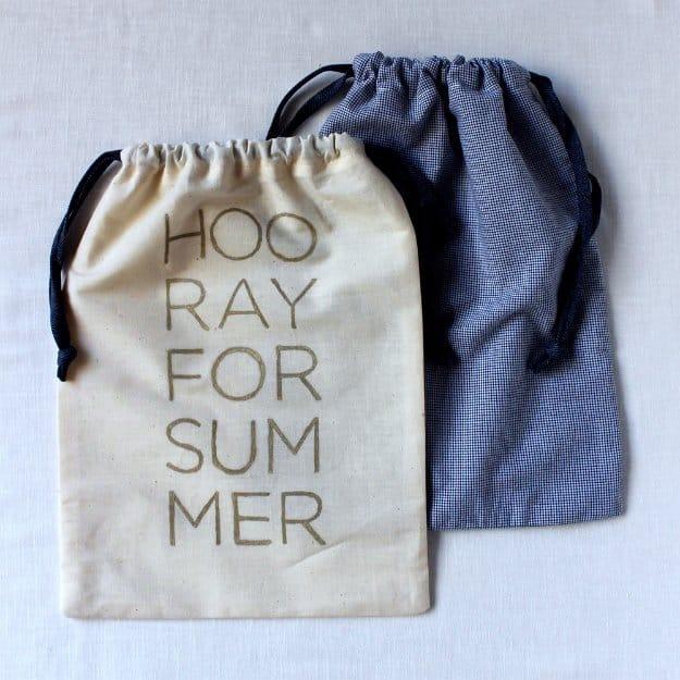A drawstring travel bag in white woven cotton lettered HOORAY FOR SUMMER in gold, tied with ribbon beside a packed case.