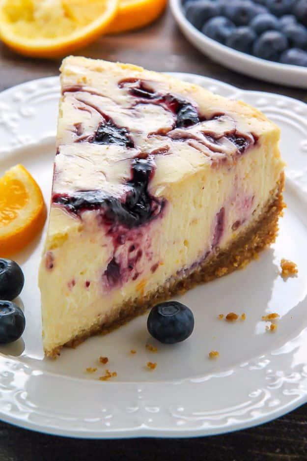 Lemon blueberry swirl cheesecake slice with purple swirls through pale filling, fresh blueberries and orange beside
