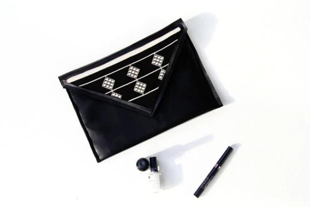 A black faux leather clutch with a zipper closure, photographed flat on a marble surface beside sunglasses.