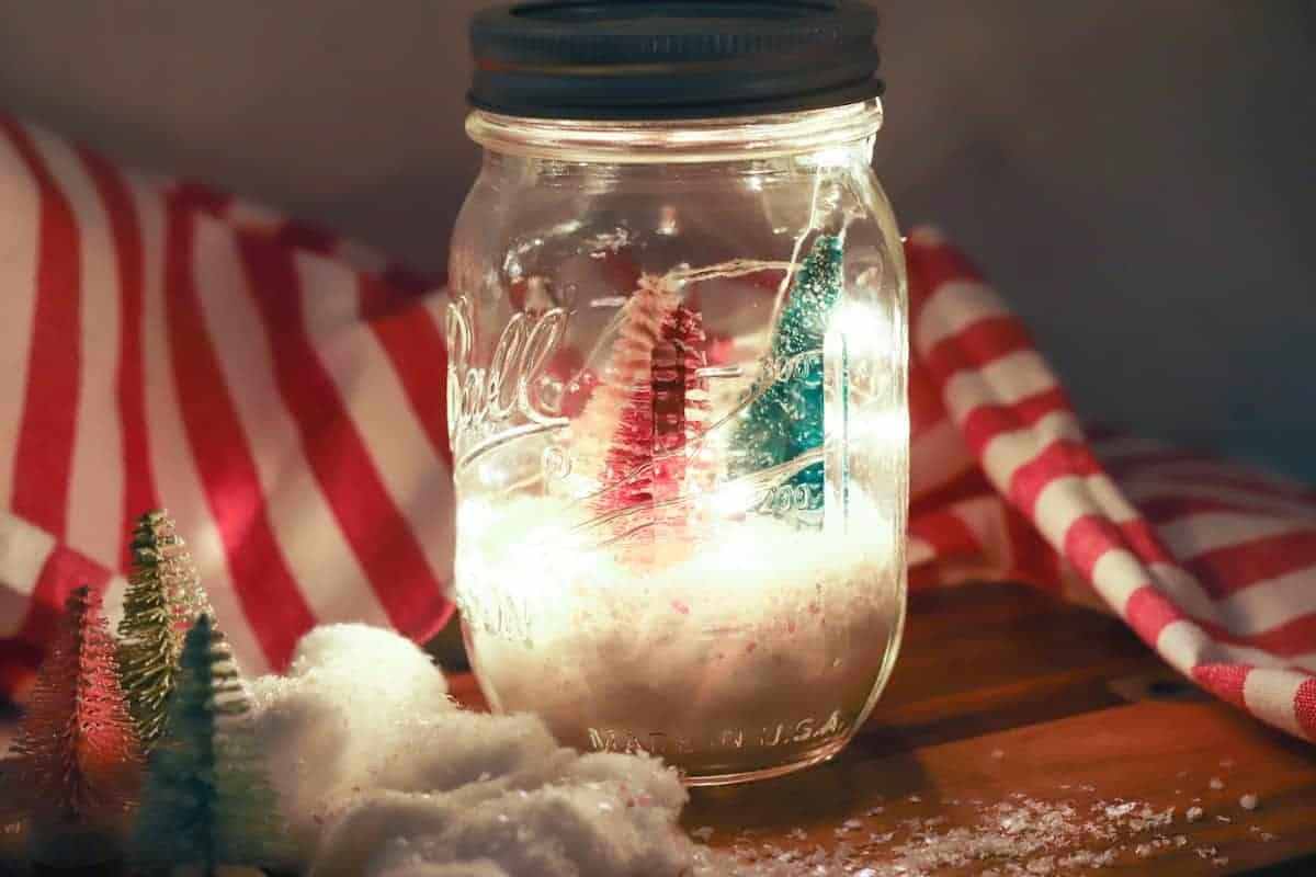 A mason jar lantern with pink and green bottle brush trees in faux snow glowing with fairy lights on a striped cloth