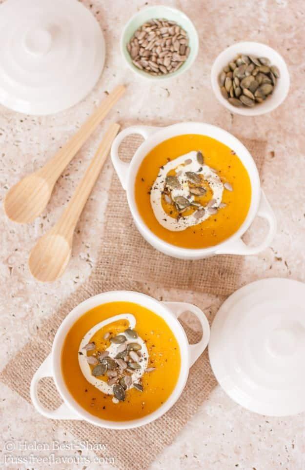 Two small white lidded pots of smooth orange pumpkin soup with cream swirls and seeds, wooden spoons