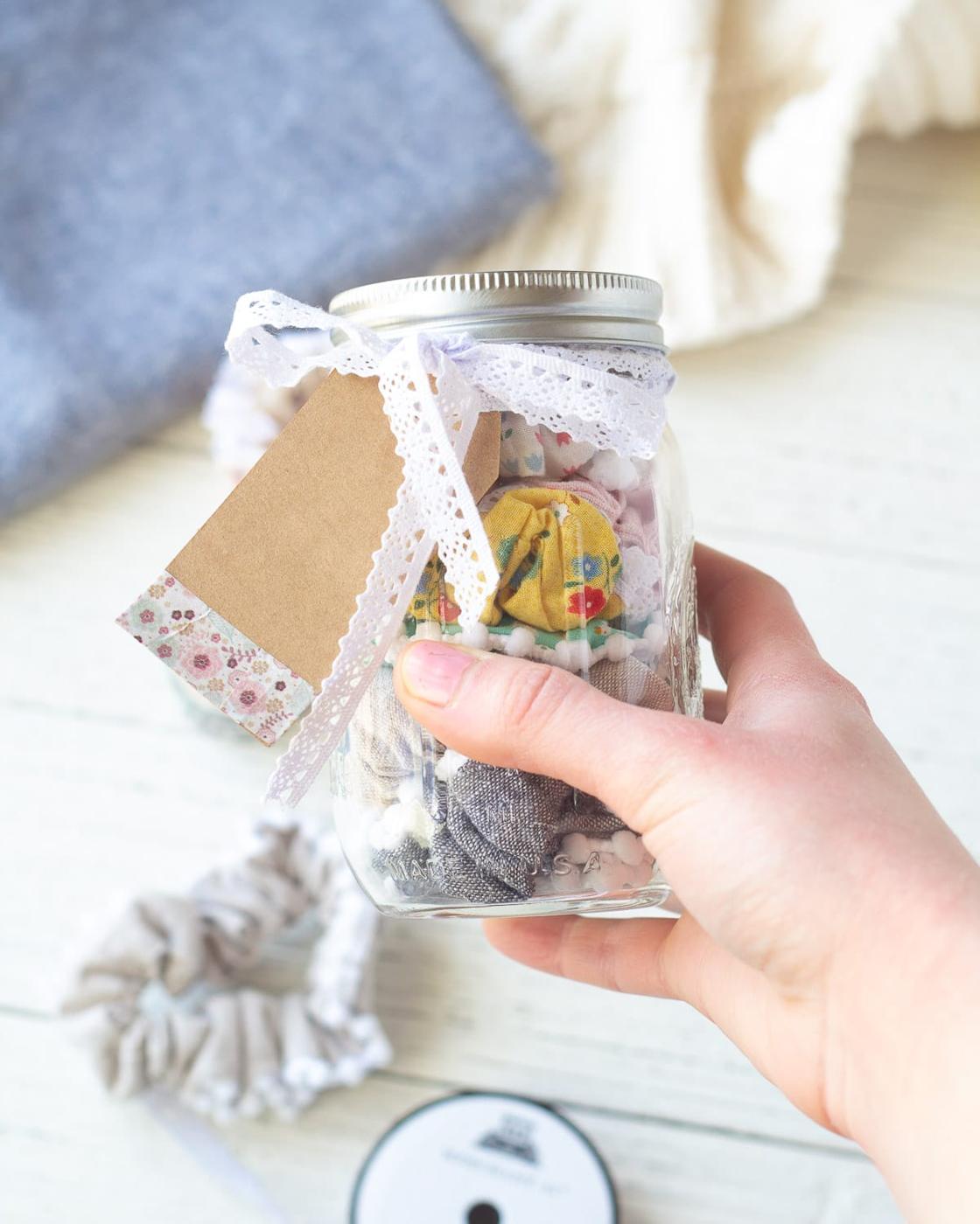 A hand holding a mason jar of handmade fabric scrunchies in mixed prints, tied with white lace ribbon and a kraft tag