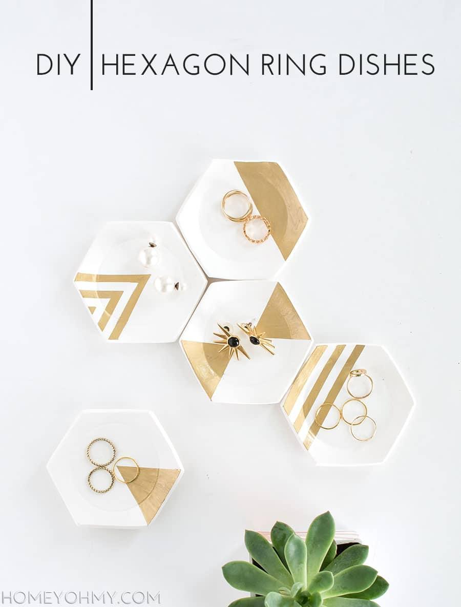 Five white hexagon clay dishes with gold painted geometric sections, holding rings and earrings beside a succulent