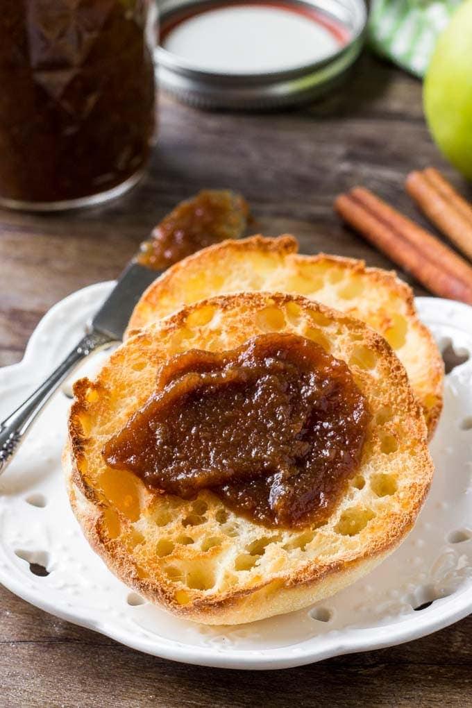 Toasted English muffin spread with dark apple butter on a white plate, with a jar of apple butter and cinnamon sticks behind