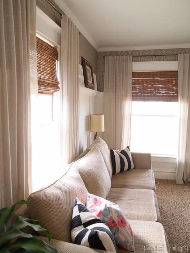 DIY window treatment of bamboo shades cut to fit, hung under cream curtain panels in a narrow living room with a neutral sofa