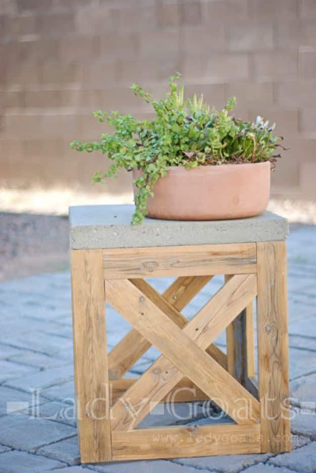 DIY X stool or side table in bare wood with X detail sides and a concrete paver top holding a pot of trailing flowers