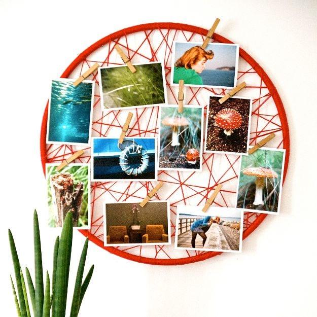 Round hoop crisscrossed with red yarn holding a dozen small photos on wooden clothespins against a white wall