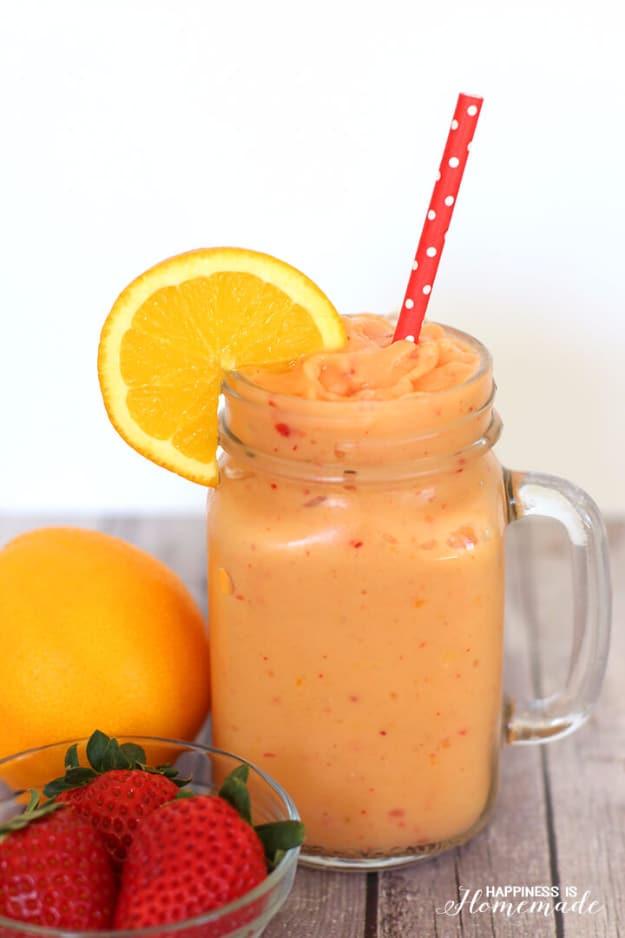 Orange coldbuster smoothie in a glass mason jar mug with a red polka dot straw, an orange slice and fresh strawberries