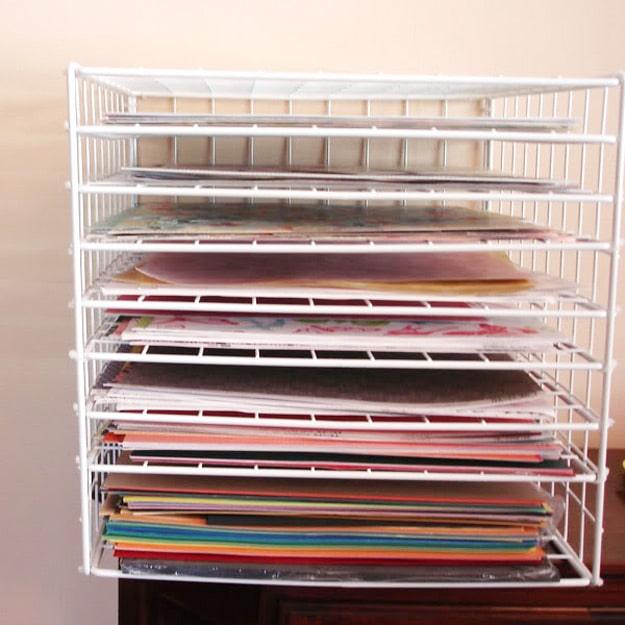White wire rack of around twenty thin shelves holding flat sheets of twelve inch scrapbook paper by color